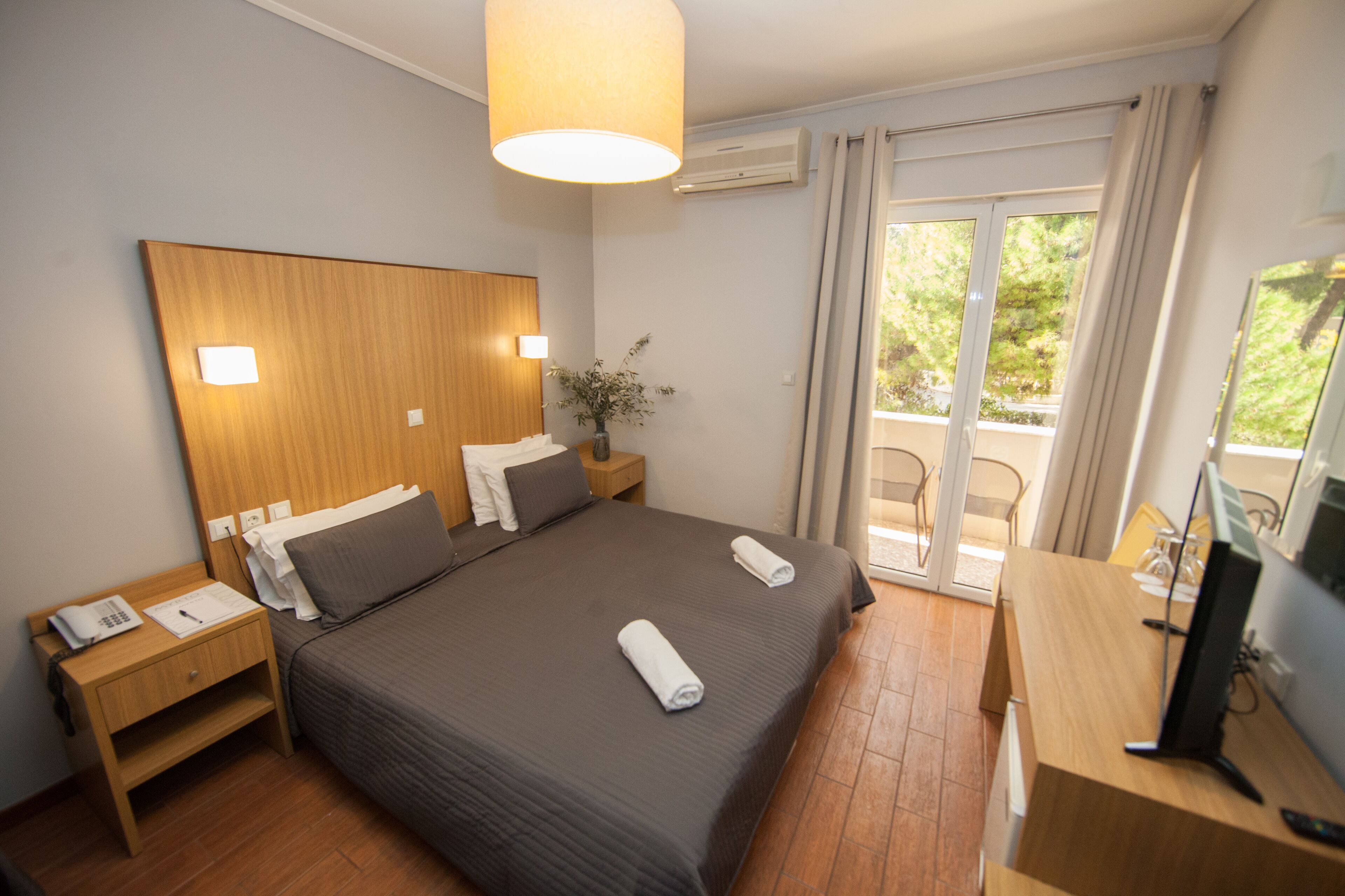 Une chambre d’hôtel avec un grand lit, une tête de lit en bois, une table de chevet avec une lampe, un bureau avec un ordinateur et une porte-fenêtre coulissante donnant sur un balcon.