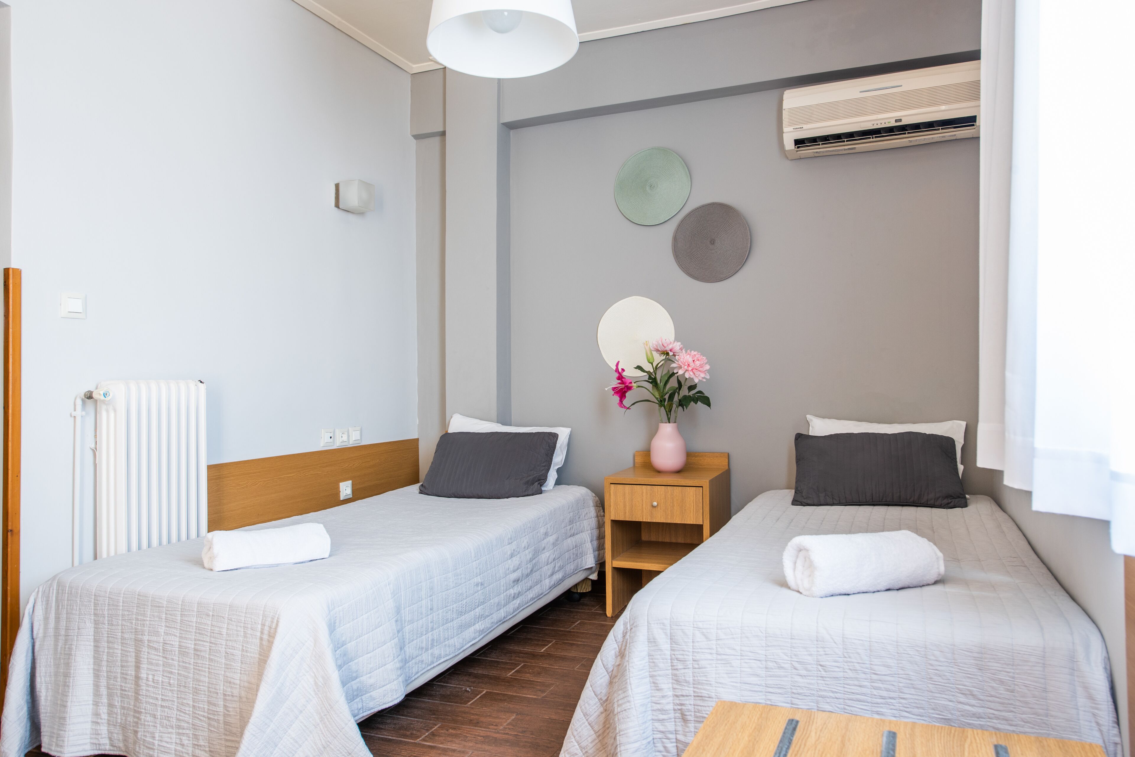 A hotel room with two single beds, a wooden nightstand, a small table, a radiator, and an air conditioning unit on the wall.