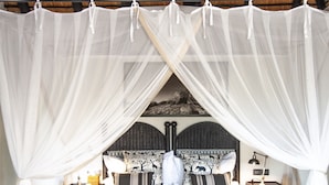 A bedroom with a bed under a canopy, a nightstand with a lamp, and a leather ottoman with books. - Jock Safari Lodge (Mbombela)