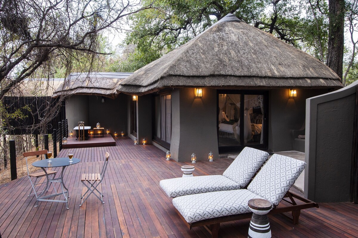 A thatched-roof outdoor seating area with lounge chairs and a table, surrounded by a wooden deck and trees.