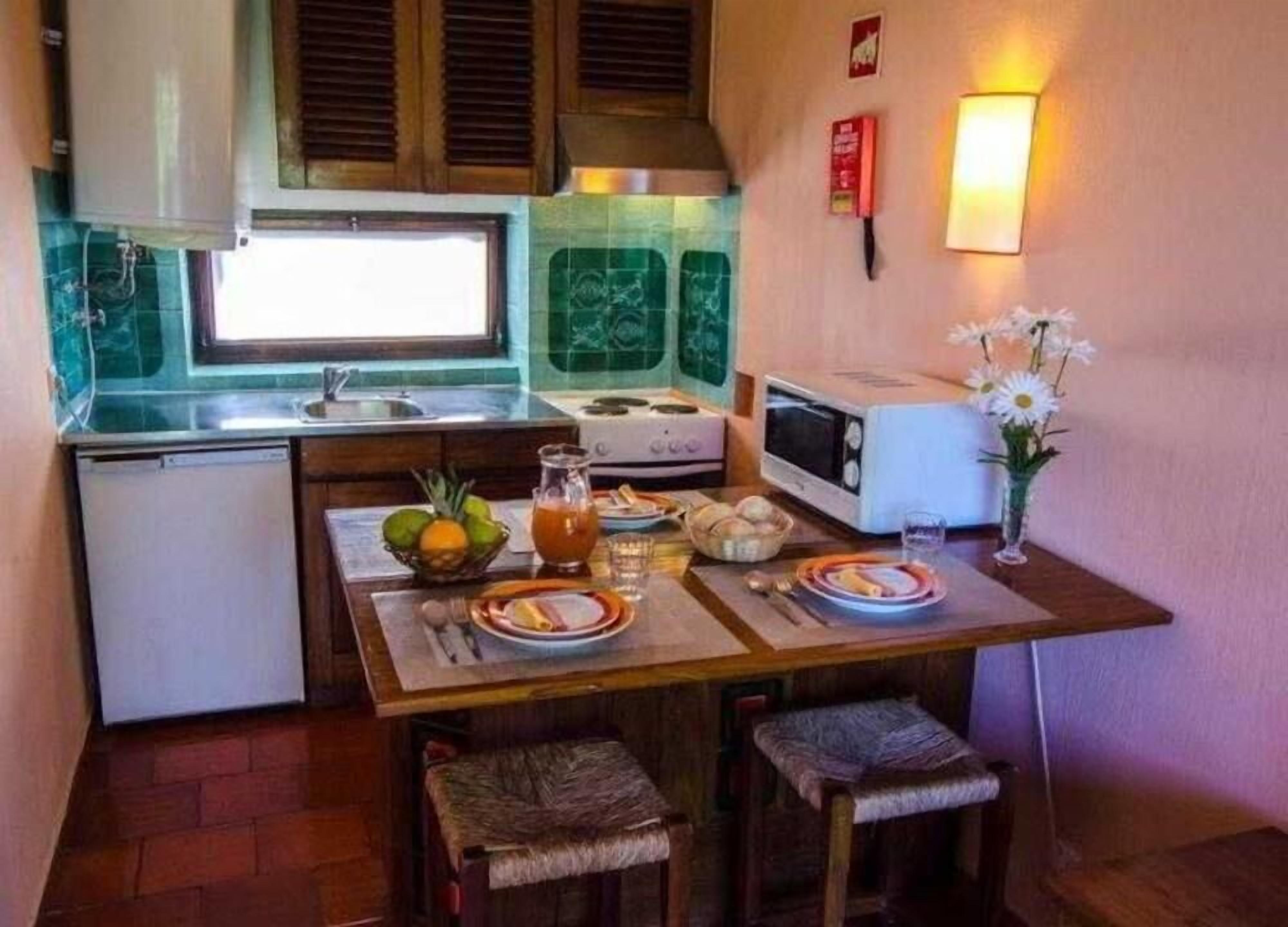 A compact kitchen with a refrigerator, stove, microwave, and a small sink. There is a dining table with plates, a pitcher, and a vase with flowers.