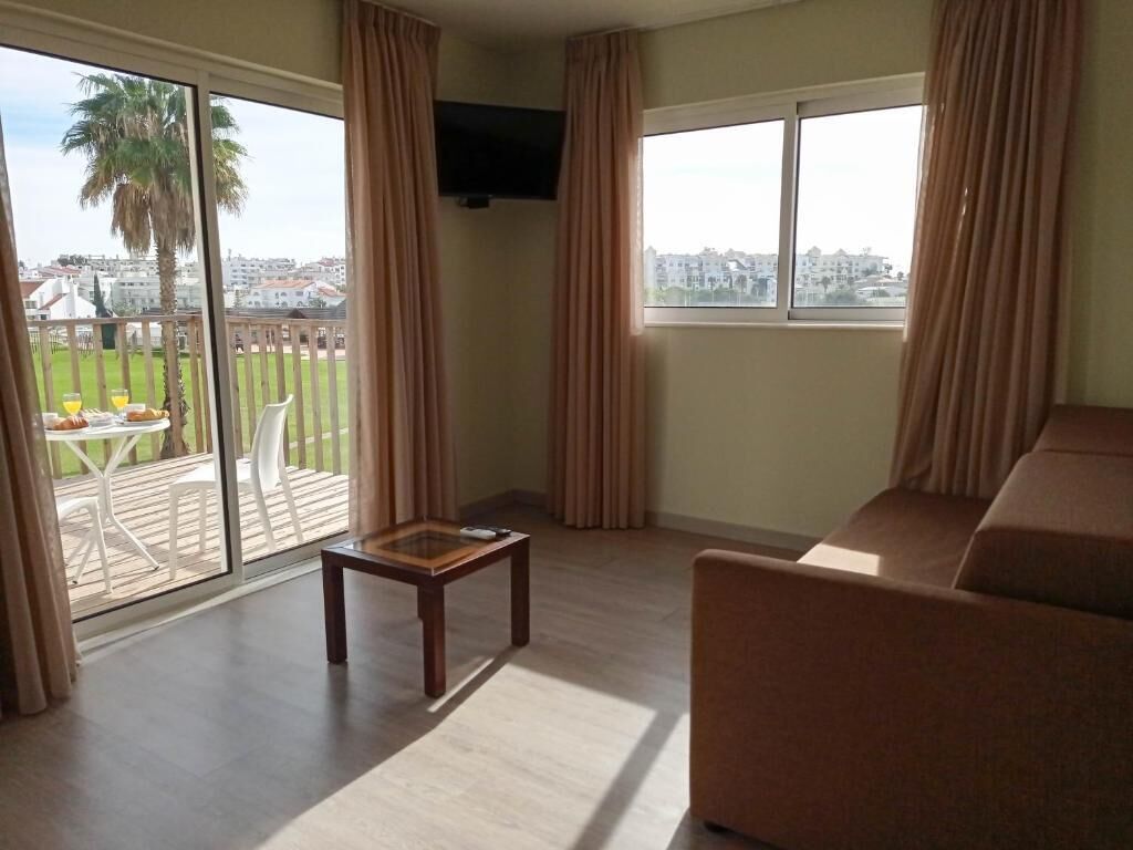 A living room with a sofa, a small table, and a TV. There is a balcony with a table and chairs, and a view of buildings and palm trees.