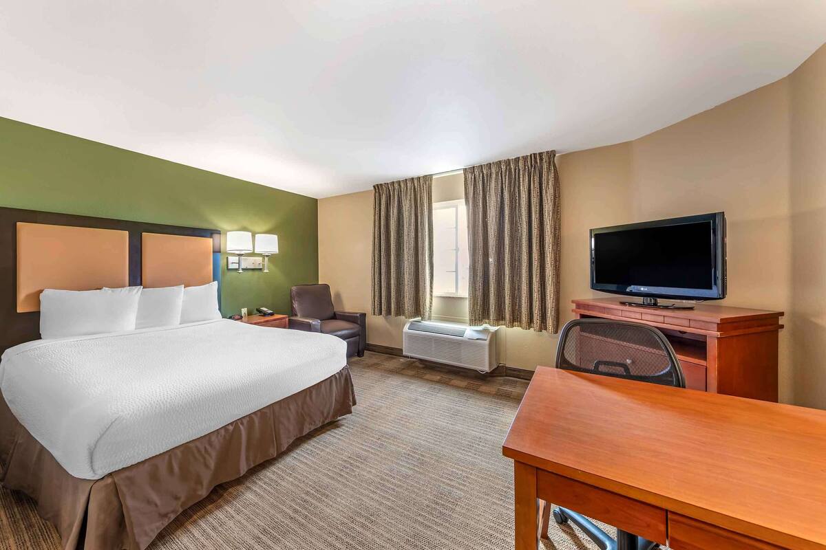 Une chambre d’hôtel avec un grand lit, un bureau, une télévision, une chaise et une fenêtre avec des rideaux.