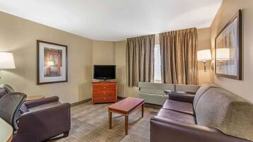 A hotel room with a sofa, armchair, TV, wooden table, and a large window with curtains.