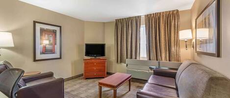 Habitación de hotel con sofá, sillón, televisor, mesa de madera y un gran ventanal con cortinas.