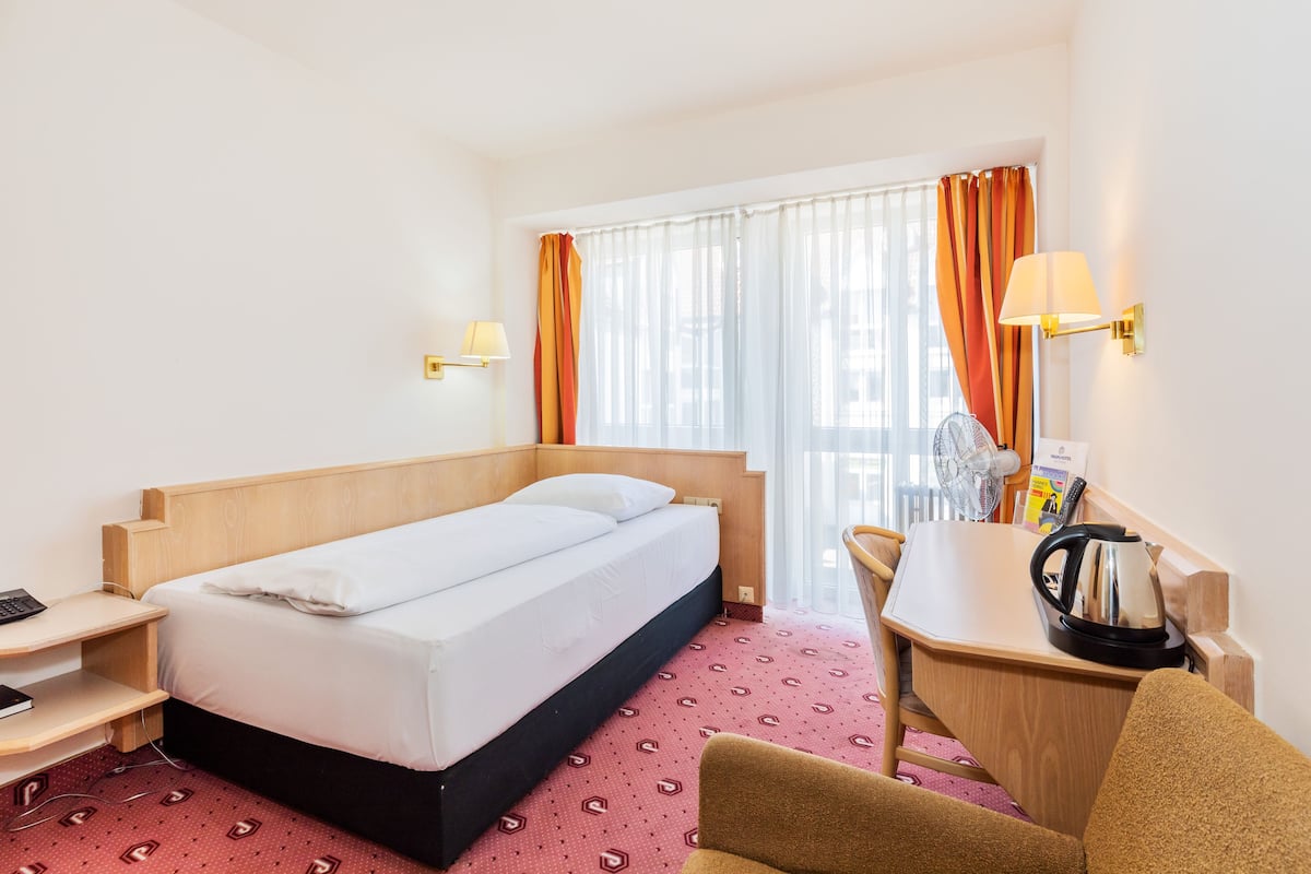 Une chambre d’hôtel avec un lit simple, un bureau avec une bouilloire, une chaise, une petite table et une fenêtre avec des rideaux.
