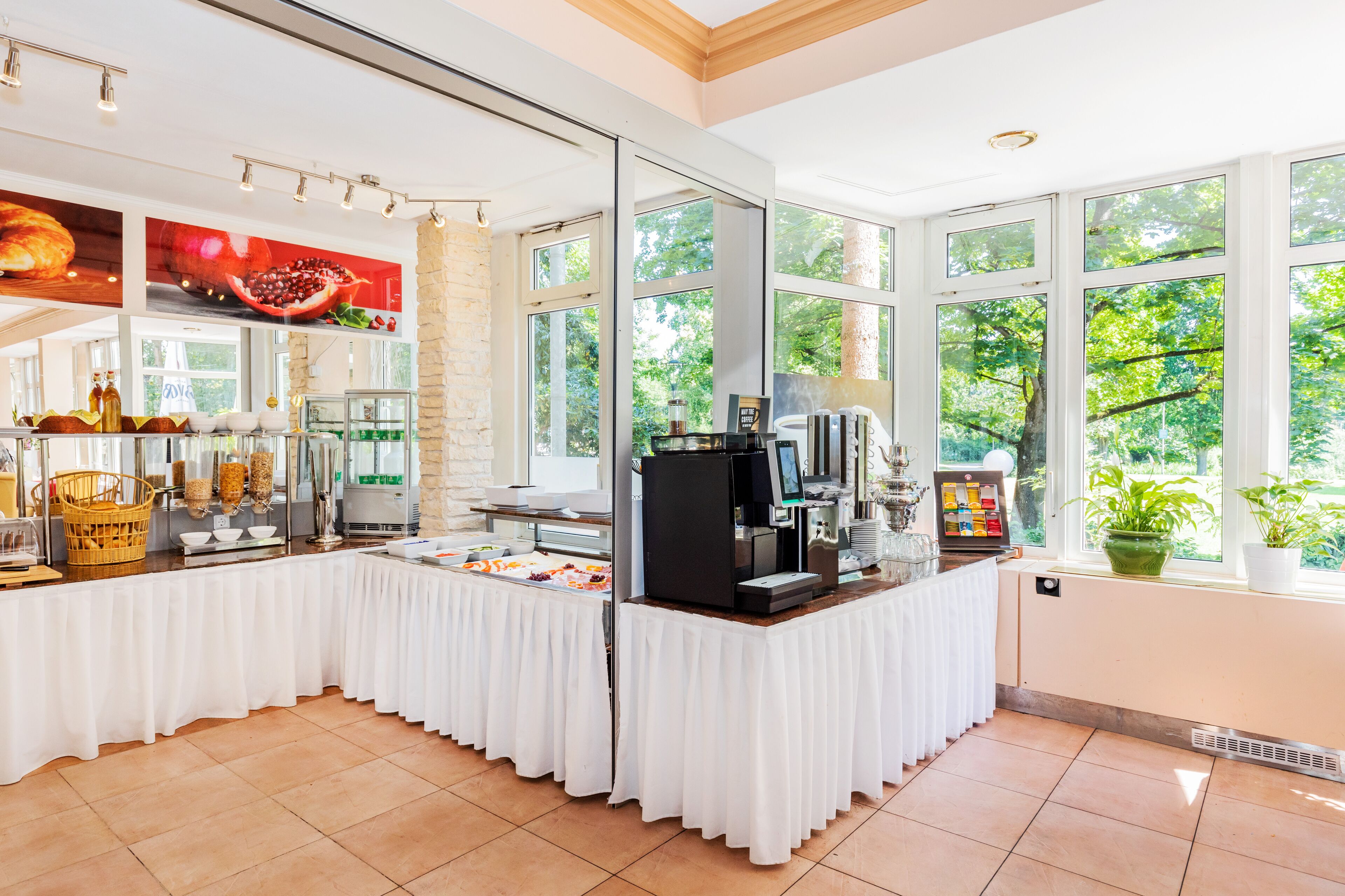 A breakfast buffet area with a coffee machine, food display, and large windows offering a view of greenery.