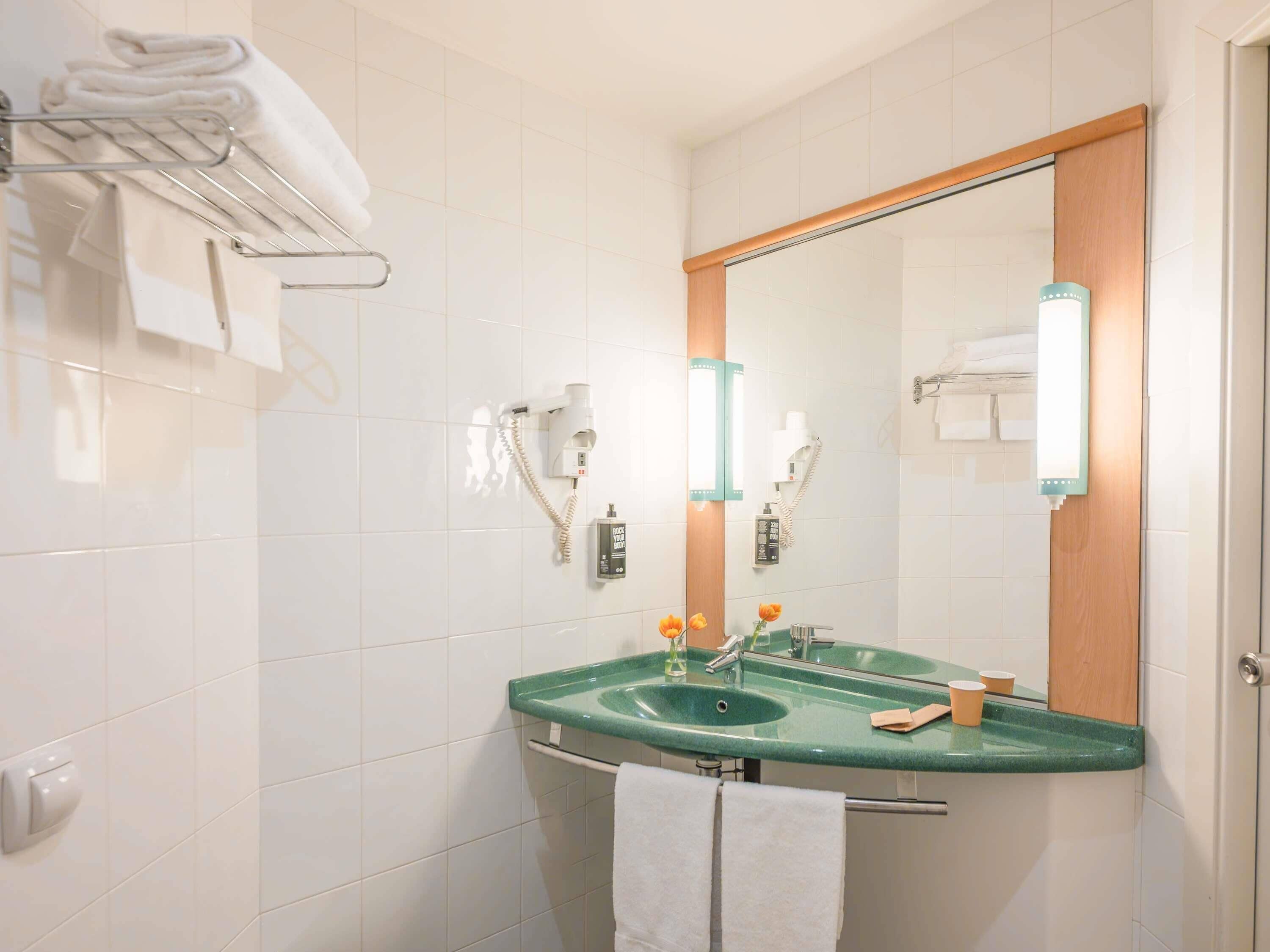 A modern bathroom with white tiles, a green sink, a mirror with wooden trim, and a towel rack with folded towels.