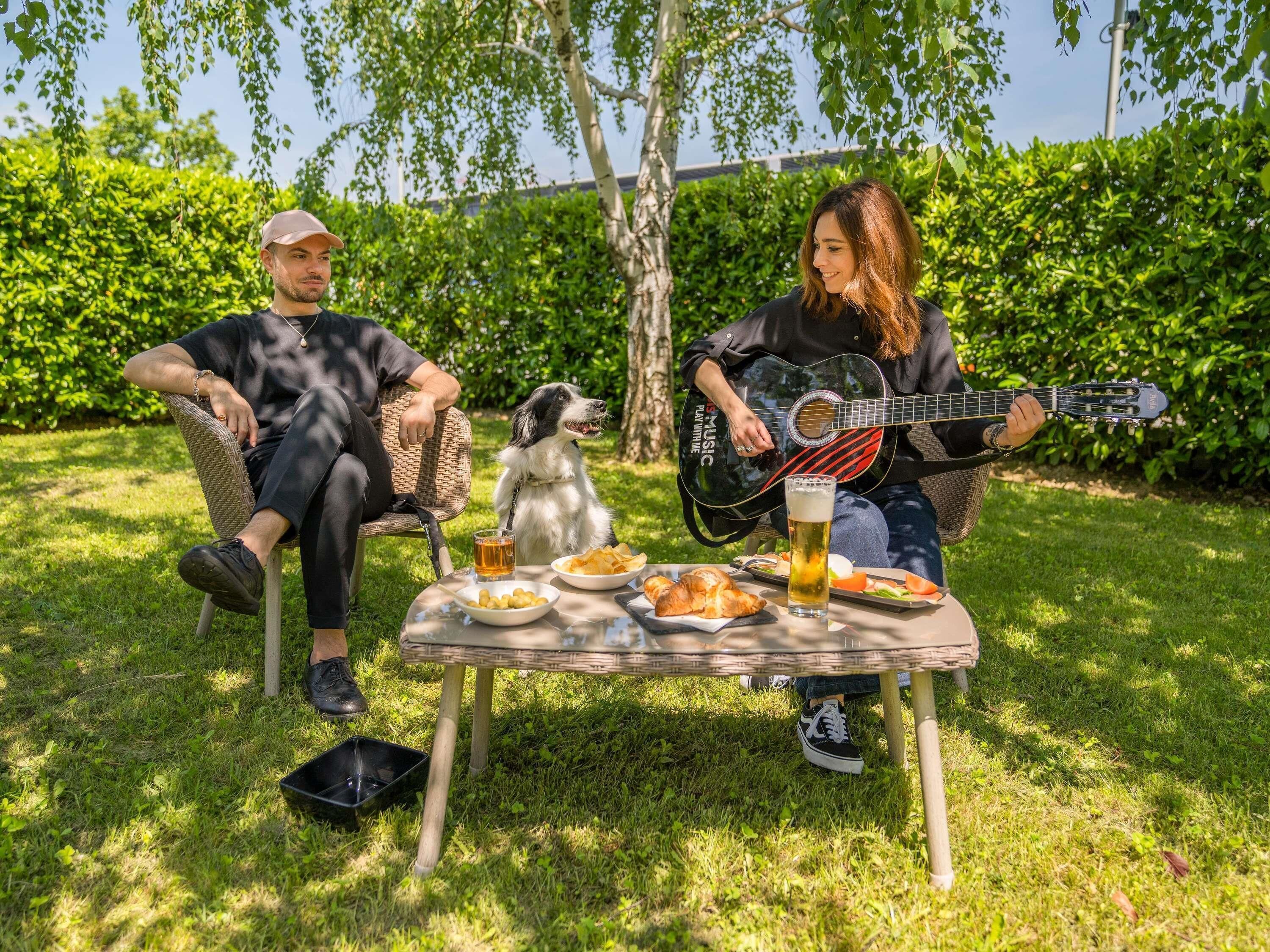 Dos personas sentadas en sillas de mimbre al aire libre, con un perro, una guitarra y comida en una mesa.