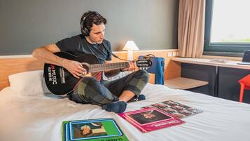 A person sitting on a bed playing a guitar, with a lamp and a suitcase in the background.