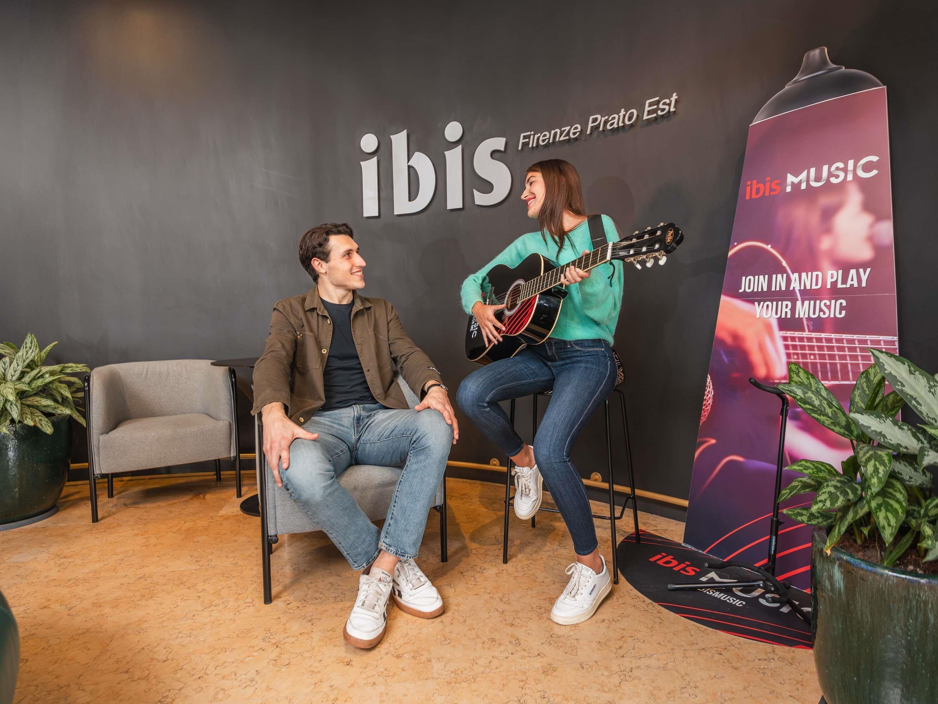 Deux personnes sont assises sur des chaises, l’une joue de la guitare. On peut voir une grande enseigne avec le mot « ibis » et une affiche qui invite à participer et à jouer de la musique.
