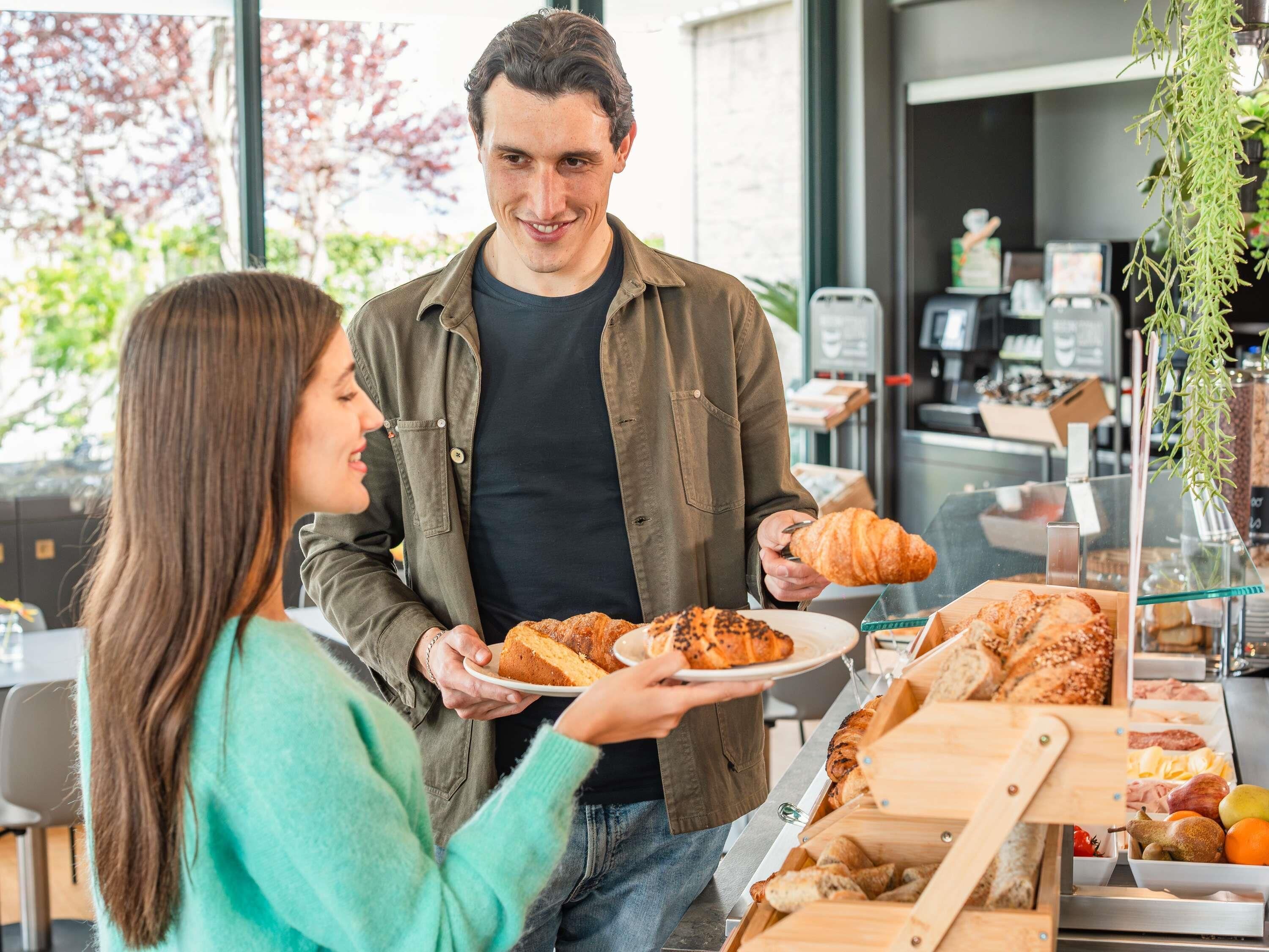 A person holding a plate with food items and a croissant, standing in front of a counter with various food items and a plant.