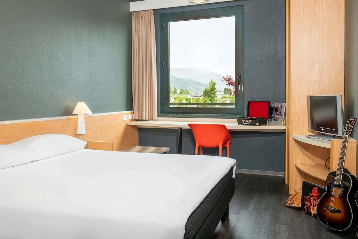 Une chambre d’hôtel équipée d’un lit, d’un bureau, d’une chaise, d’une télévision, d’une guitare et d’une fenêtre donnant sur un paysage agréable.
