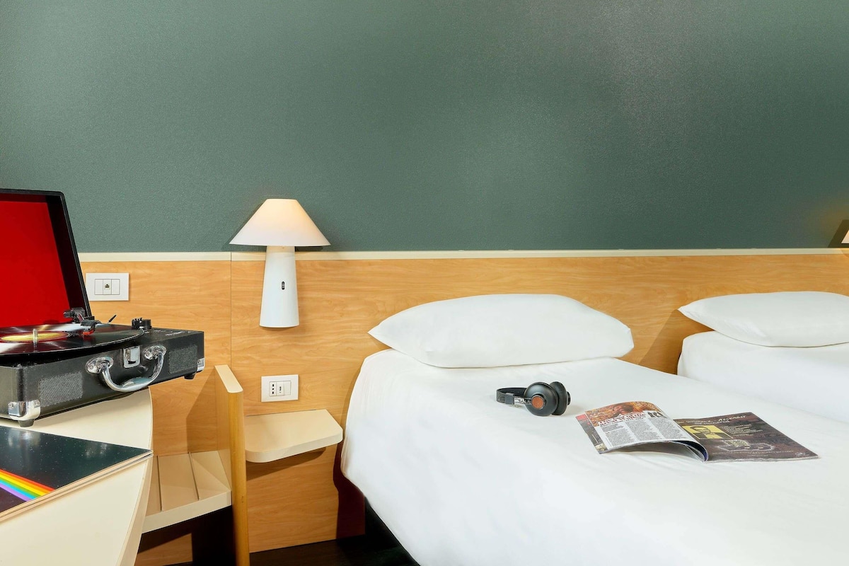 Une chambre d’hôtel avec un lit, une table de chevet avec une lampe, une valise, une paire d’écouteurs et un magazine.