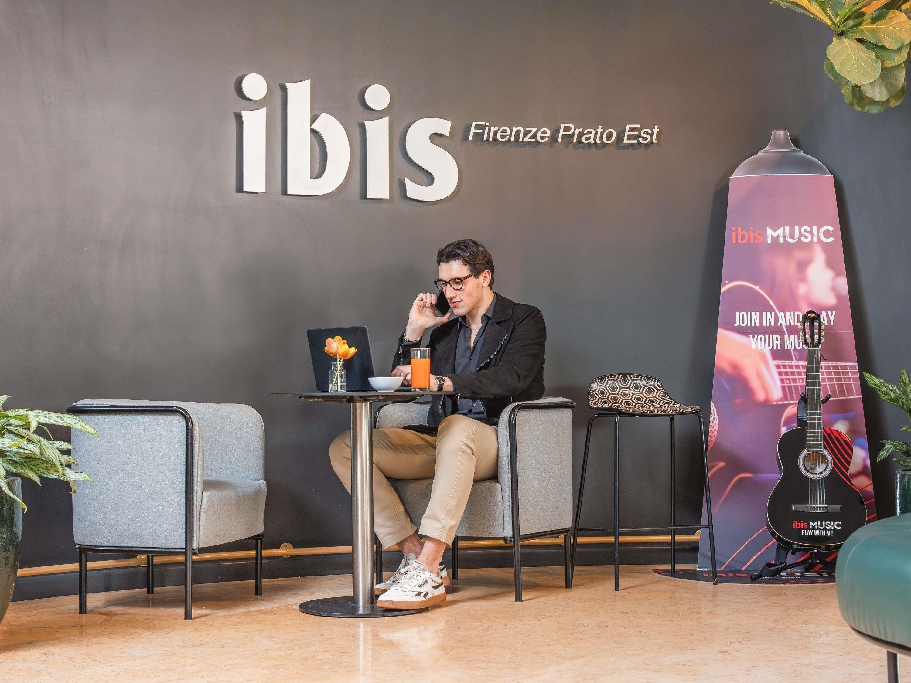 Een man zit aan een tafel met een laptop en een drankje, voor een muur met de tekst 'ibis' en een gitaar.