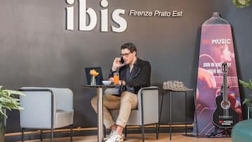 Un hombre sentado a una mesa con una computadora portátil y una bebida, frente a una pared con la palabra 'ibis' y una guitarra.