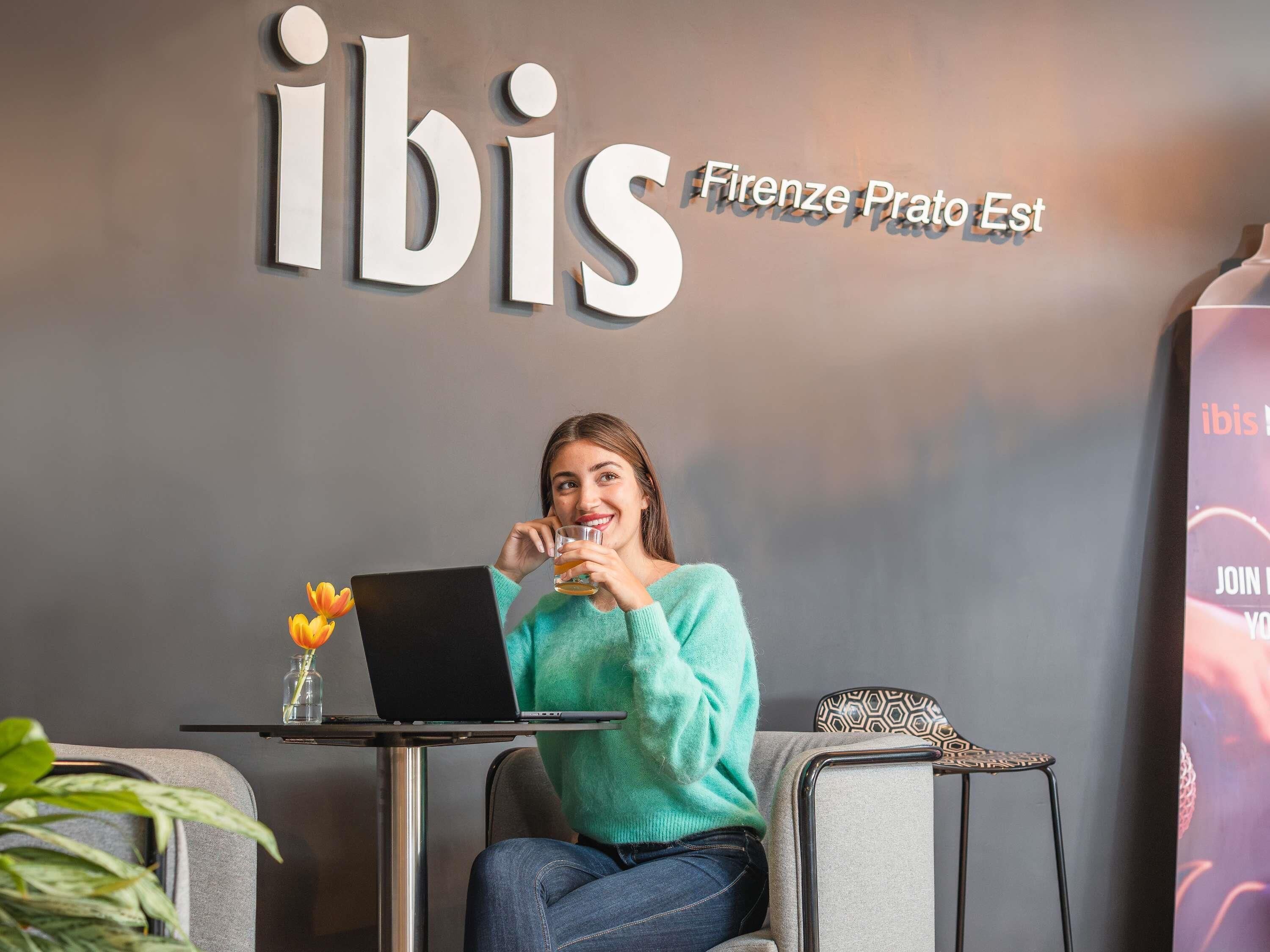 A person sitting at a table with a laptop, a glass of drink, and a vase with flowers. The wall behind has a sign with the word 'ibis' and 'Firenze Prato Est'.