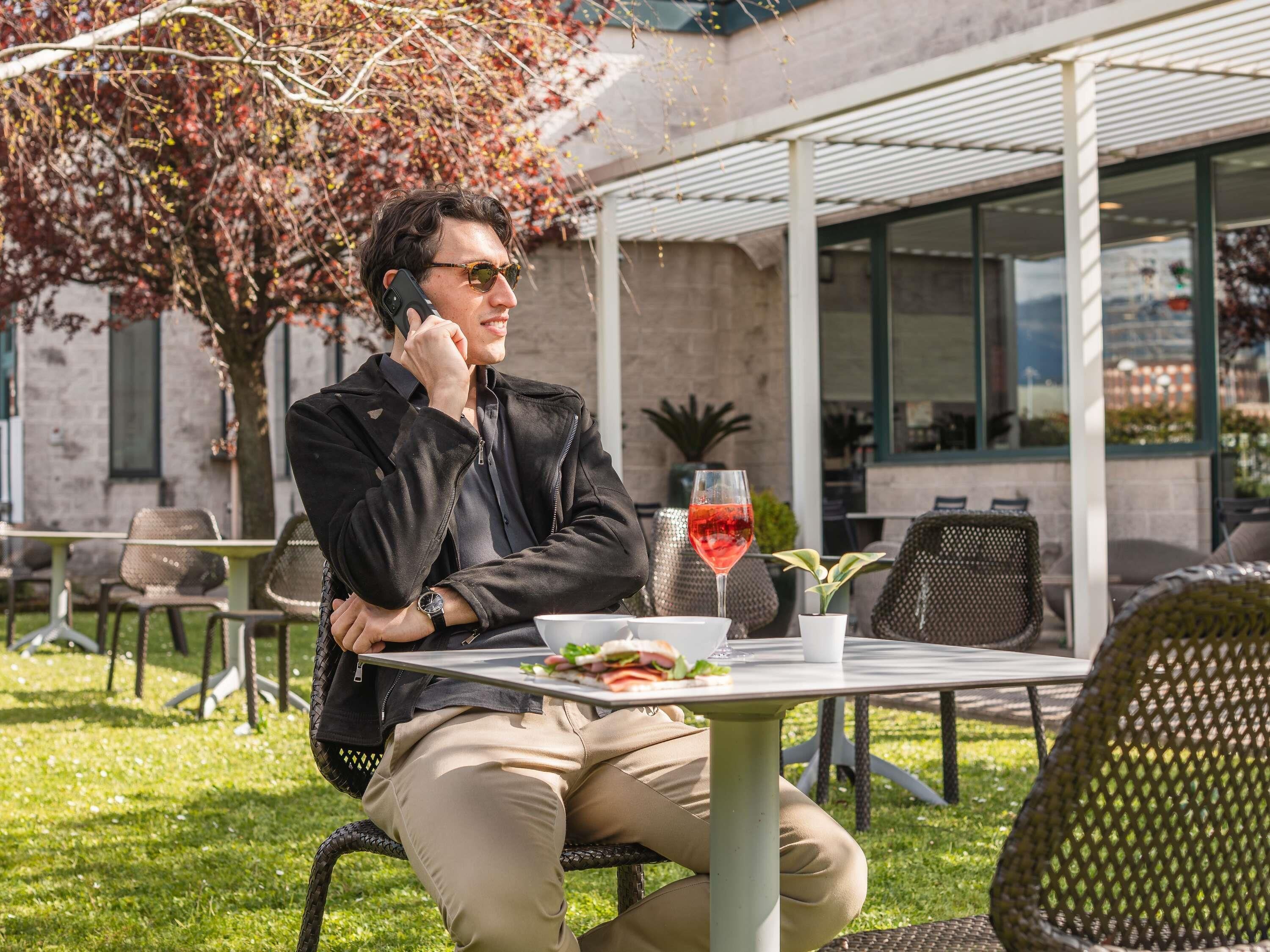 A person sitting at an outdoor table with a drink and food, in a setting with chairs and a building in the background.