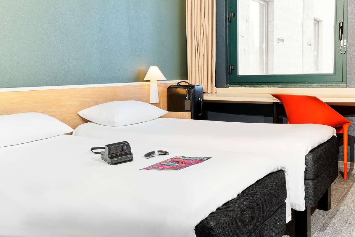A hotel room with a bed, a desk, a lamp, a suitcase, a chair, and a window with curtains.