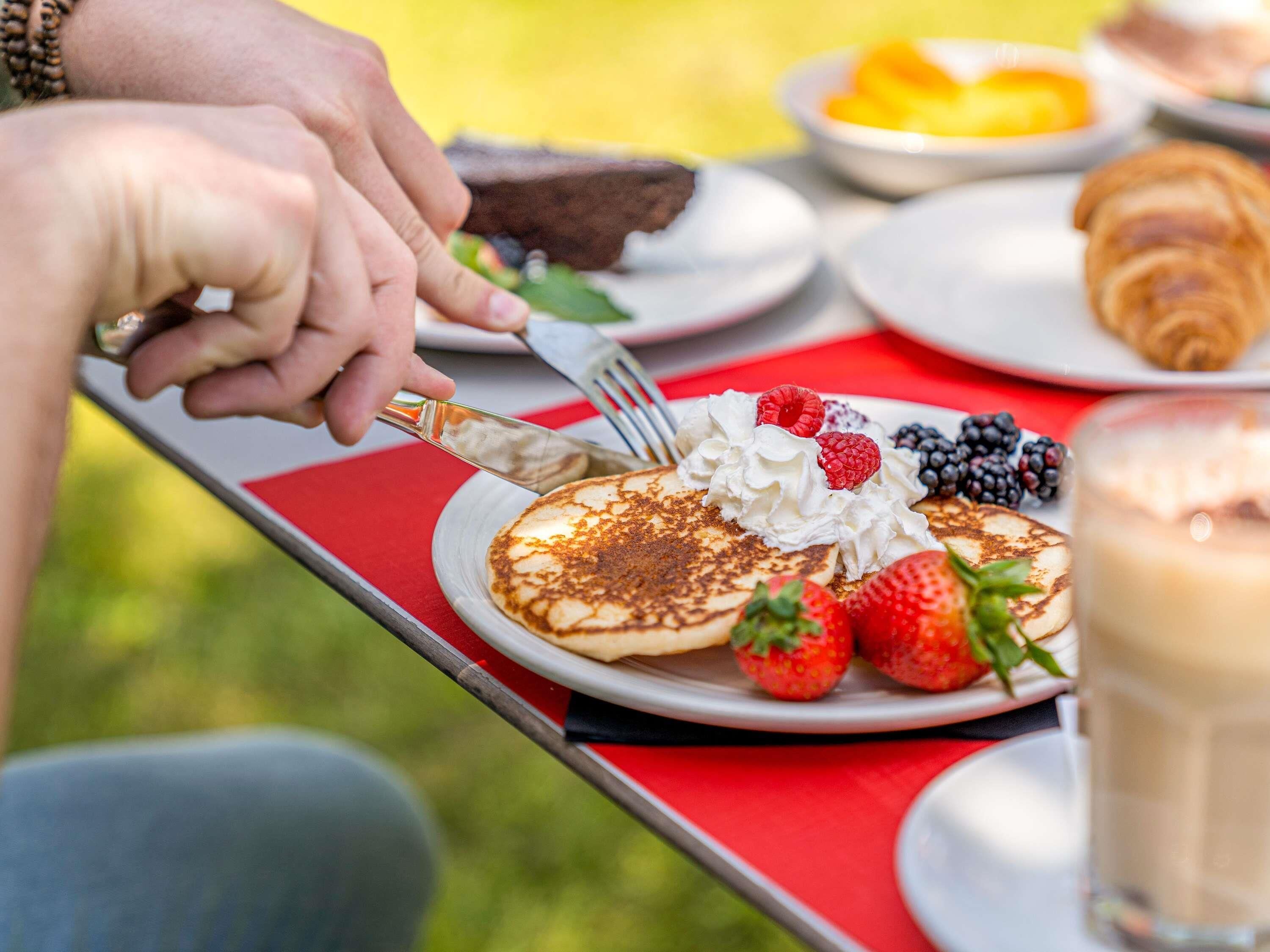 A breakfast setting with pancakes topped with whipped cream and berries, a croissant, and a glass of milk on a red tablecloth.