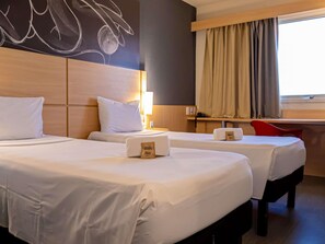 A hotel room with two beds, a desk, a chair, a wall art piece, and a window with curtains.