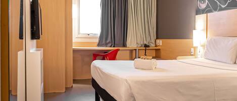 A hotel room with a bed, a desk with a chair, a TV, and a window with curtains.
