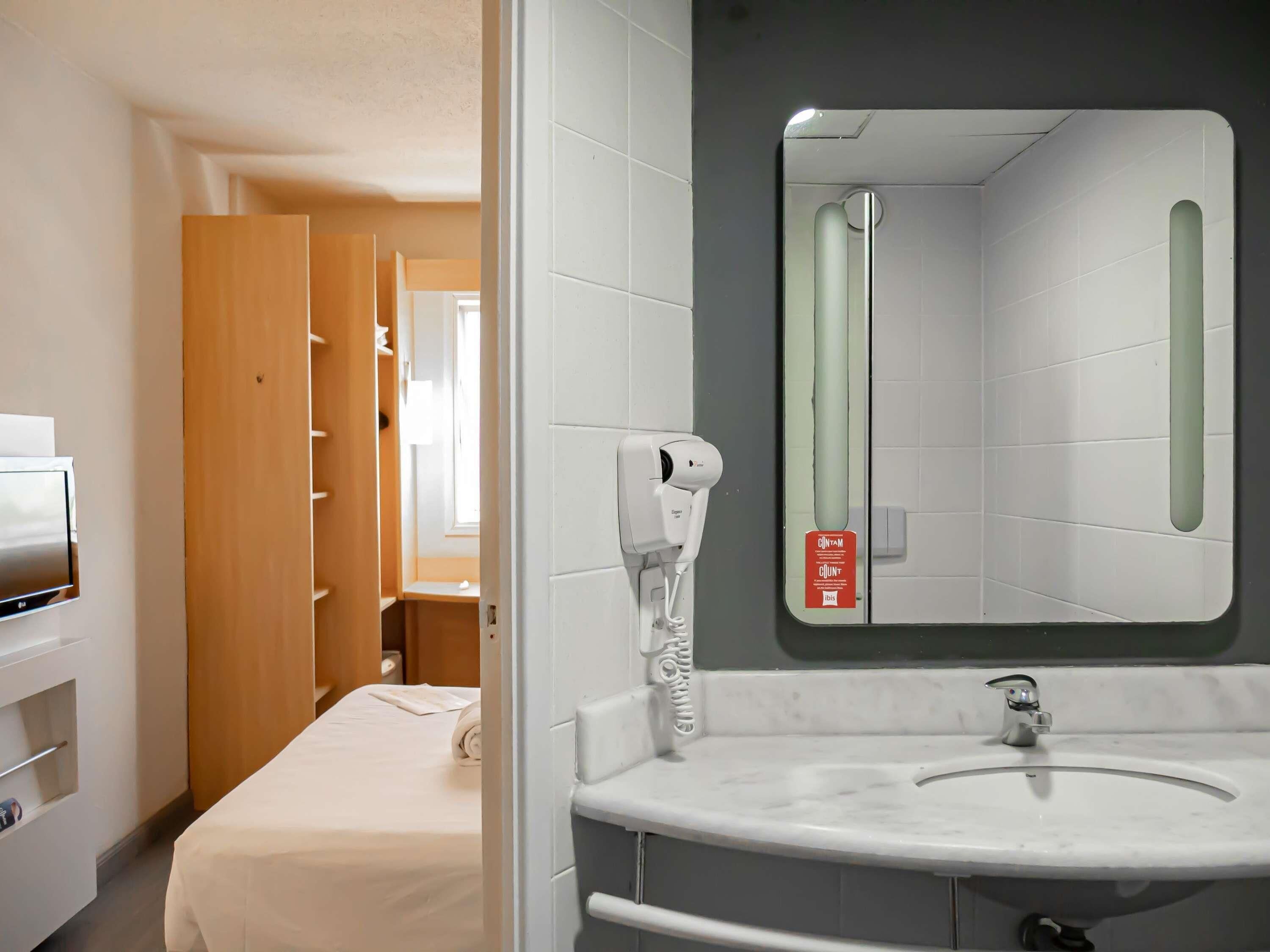A hotel room with a bed, a white bathroom with a mirror, a towel, and a hairdryer.