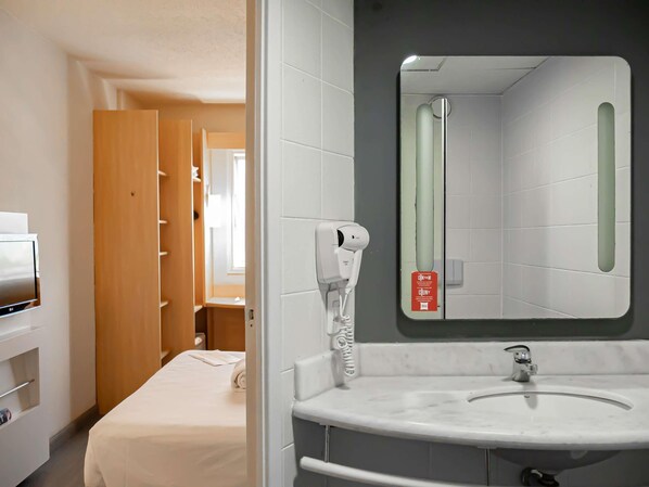 A hotel room with a bed, a white bathroom with a mirror, a towel, and a hairdryer.