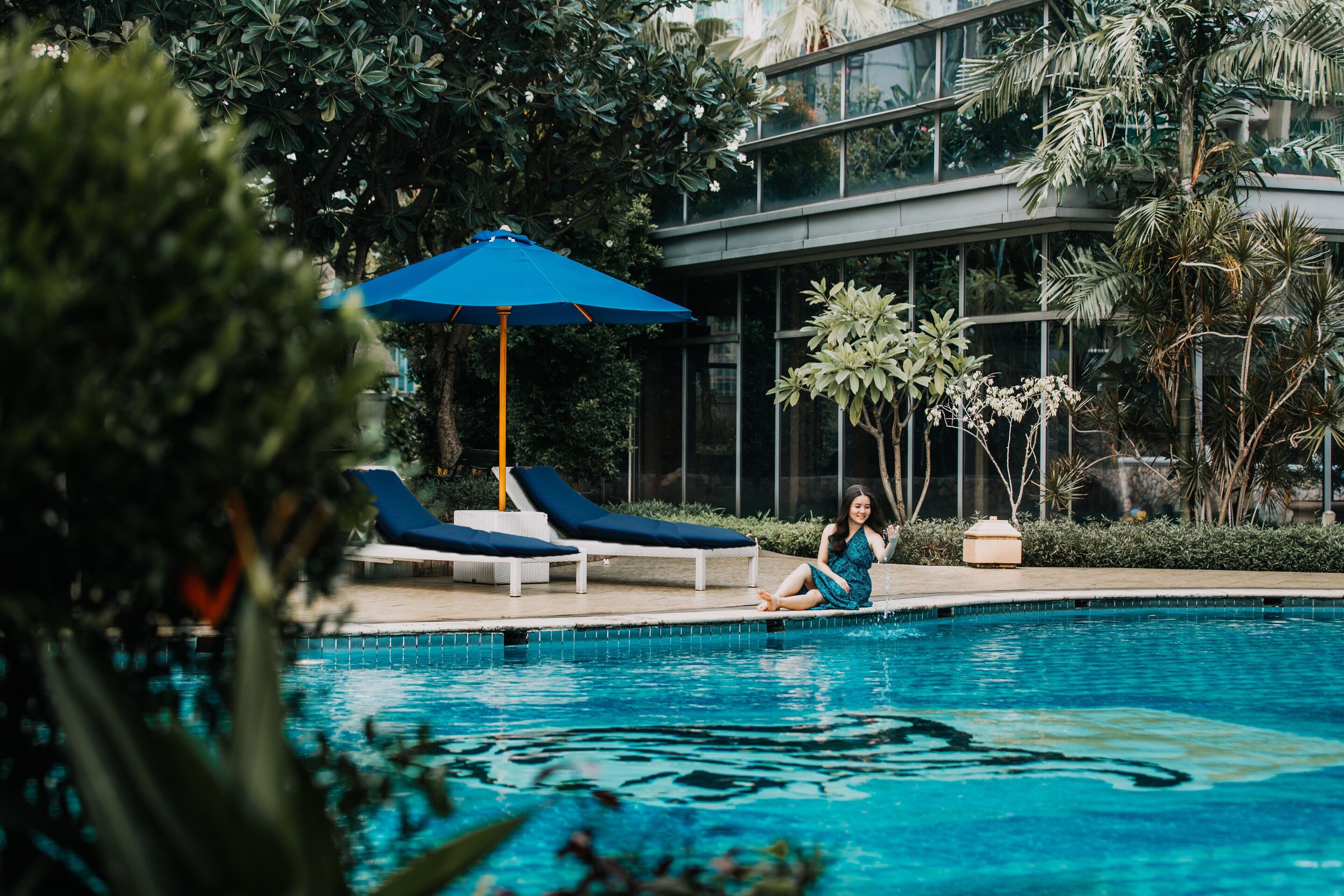 A pool area with lounge chairs and an umbrella, surrounded by lush greenery and modern architecture.