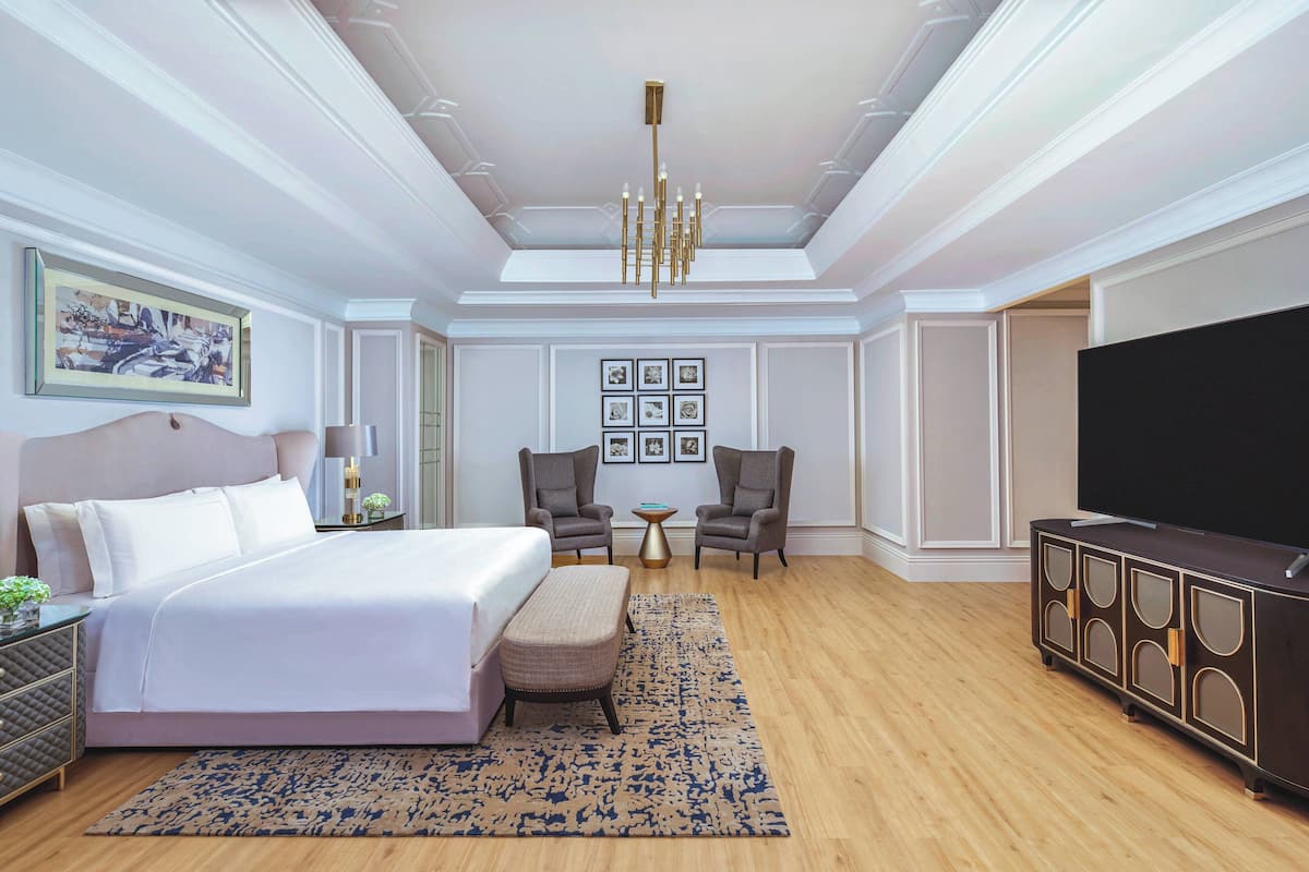 A hotel room with a large bed, two armchairs, a small table, a TV, and a chandelier.