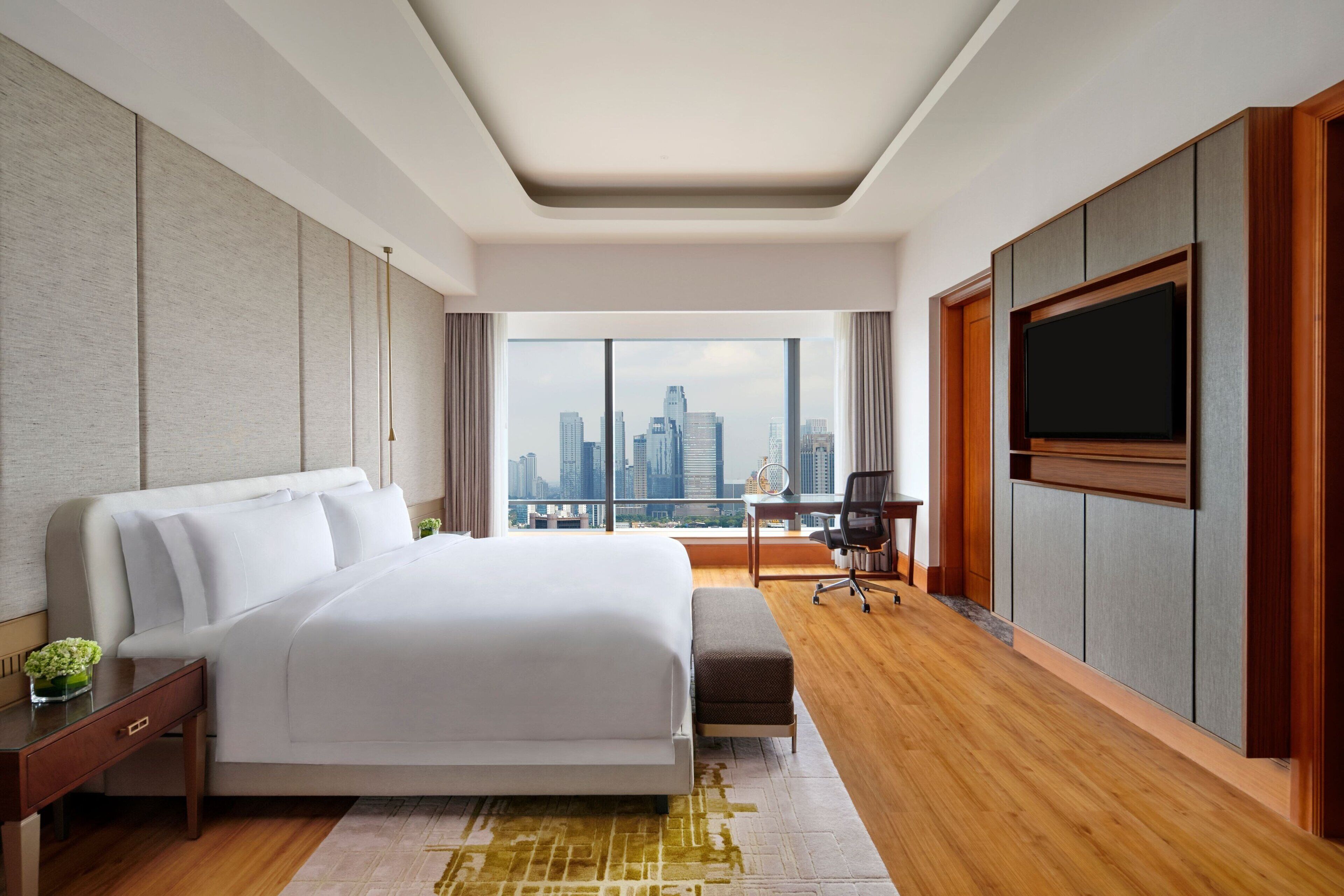 A modern hotel room with a large bed, a desk, a chair, a flat-screen TV, and a city view.