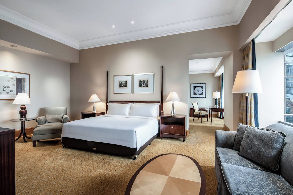 A hotel room with a large bed, two bedside tables with lamps, a grey armchair, a sofa, and a small round table.