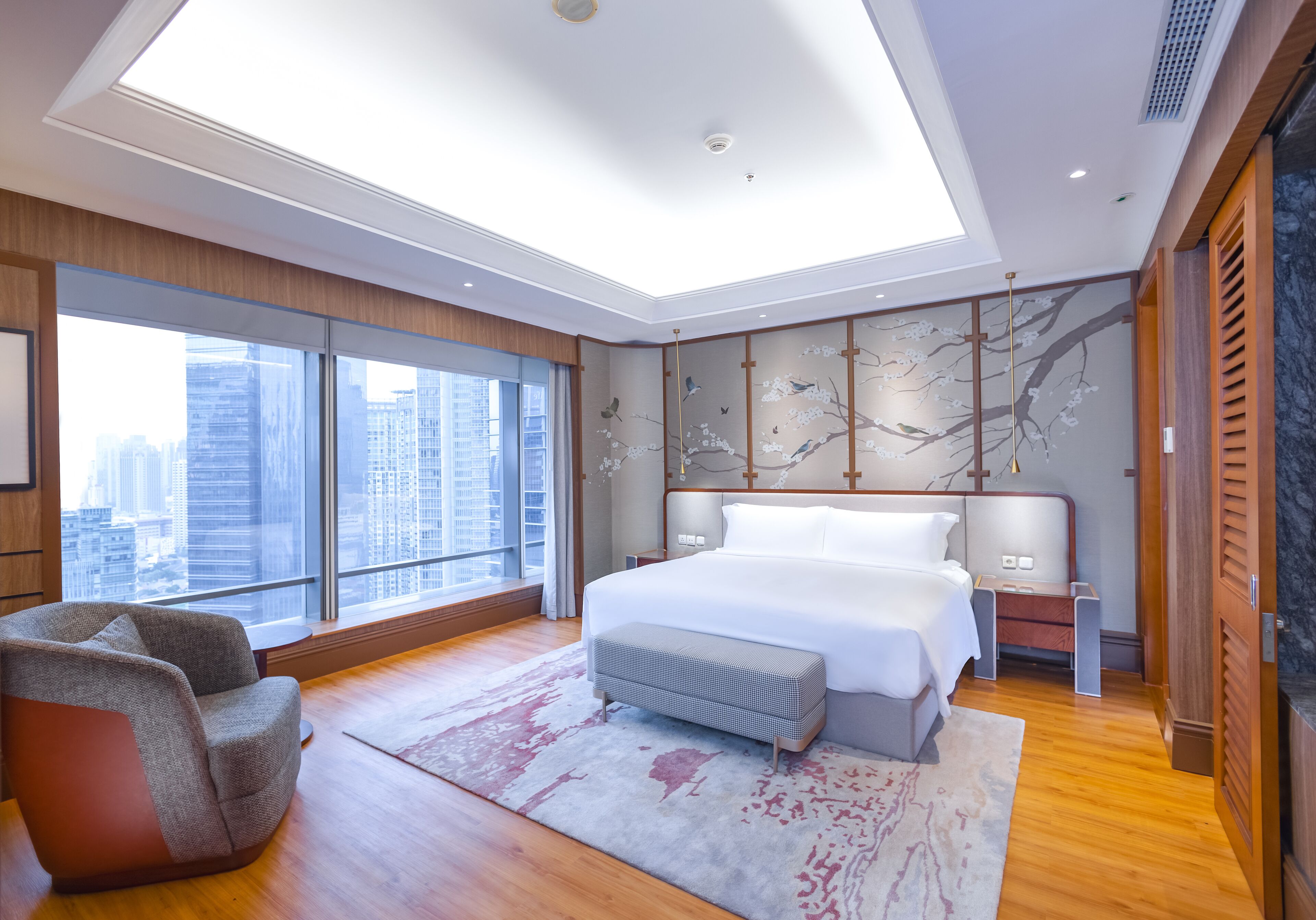 A modern bedroom with a large bed, a seating area, and a city view.