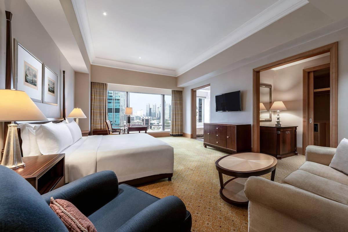 A hotel room with a large bed, two bedside tables with lamps, a sofa, a small round table, a TV mounted on the wall, and a view of the city through large windows.