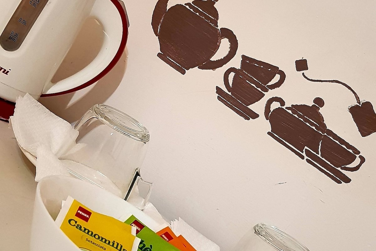 Een keukenhoek met een witte waterkoker, een theepot en een kopje op het aanrecht. Er staat een witte schaal met een Camomilla theezakje en een groen-oranje kaartje.
