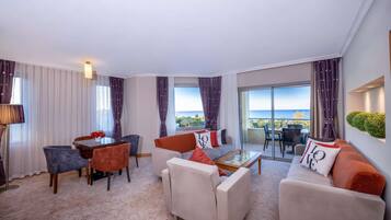 Una sala de estar moderna con sofá, sillones, una mesa de centro y una mesa de comedor. Cuenta con un ventanal con cortinas, un balcón con mobiliario exterior y vistas al mar.