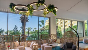 A modern lounge area with wicker furniture, a hanging chair, and large windows offering a view of palm trees and a building. - Selectum Family Resort Side (Manavgat)