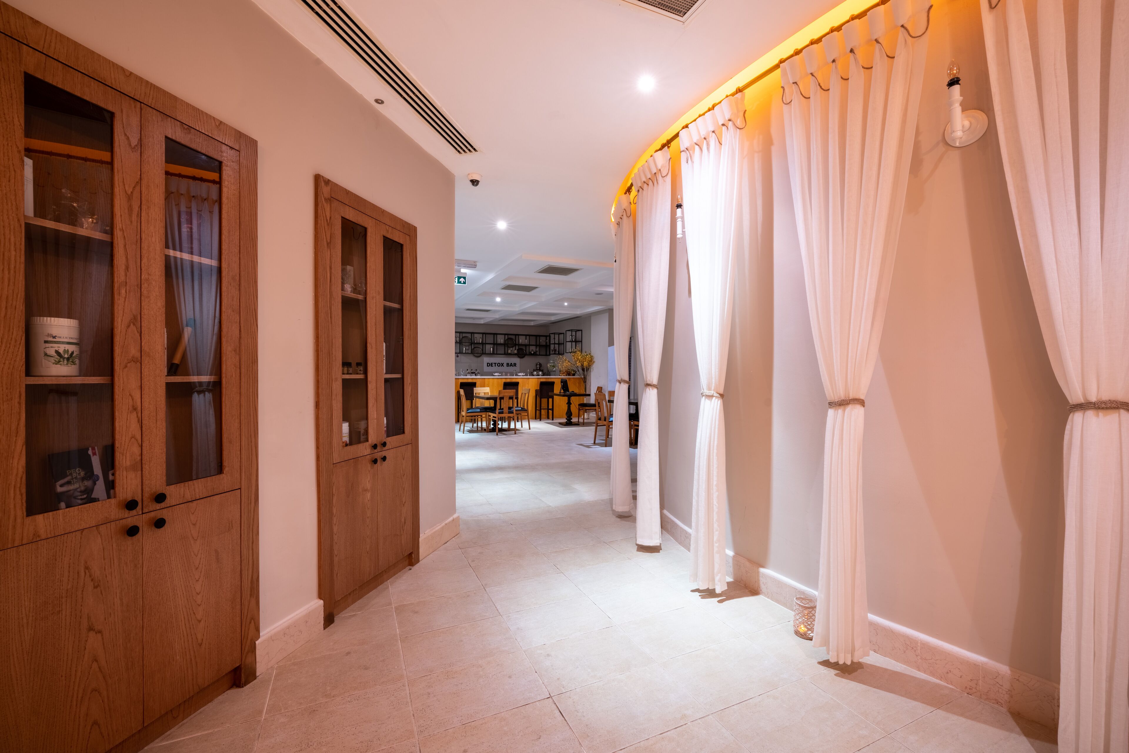 A hallway with wooden cabinets, white curtains, and a tiled floor leading to a bar area.
