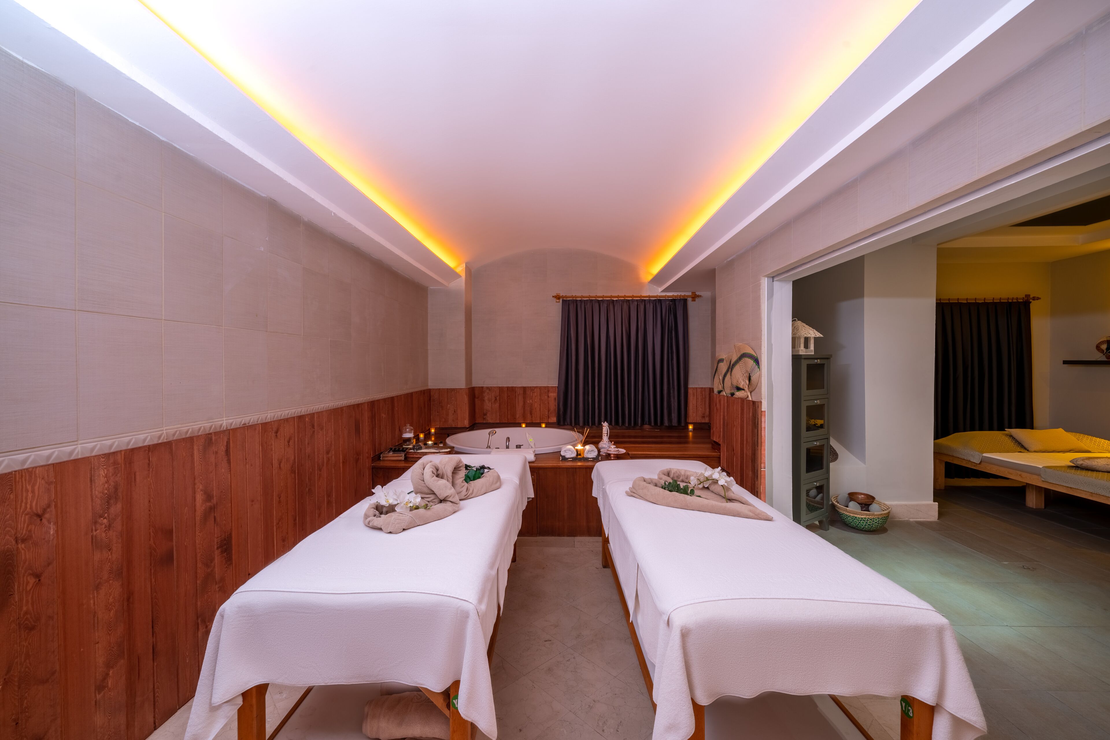 A spa room with two treatment beds, a wooden wall, a large mirror, and a small window.