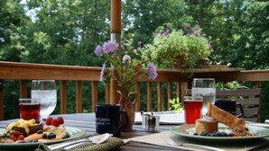 Una terraza de madera con una mesa dispuesta para el desayuno, que incluye platos de comida, bebidas y un florero con flores.