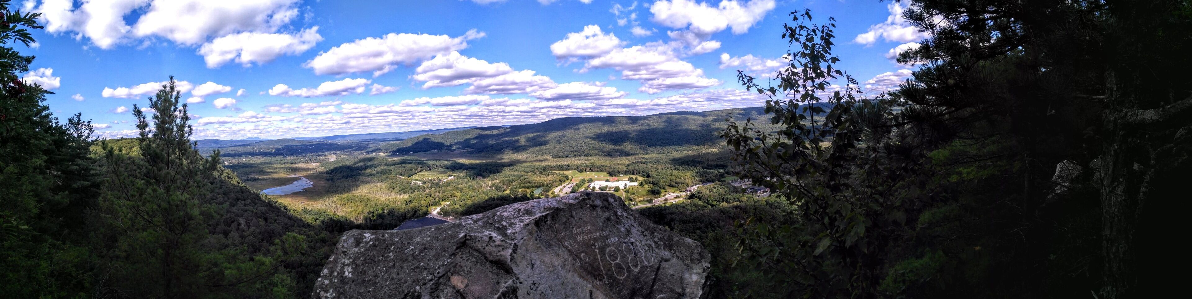A panoramic view of a valley with a house surrounded by mountains and trees.