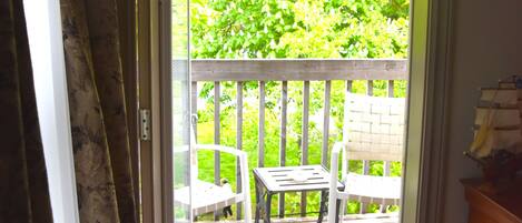 An open door leading to a wooden deck with a white chair and table, surrounded by lush greenery.