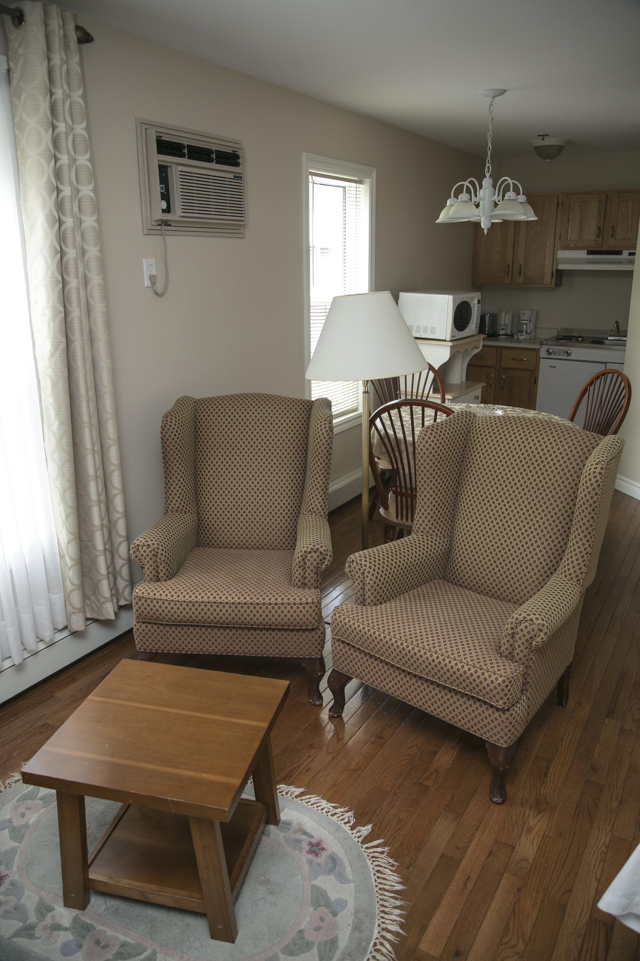 A living room with two patterned armchairs, a wooden coffee table, a lamp, and a kitchen in the background.
