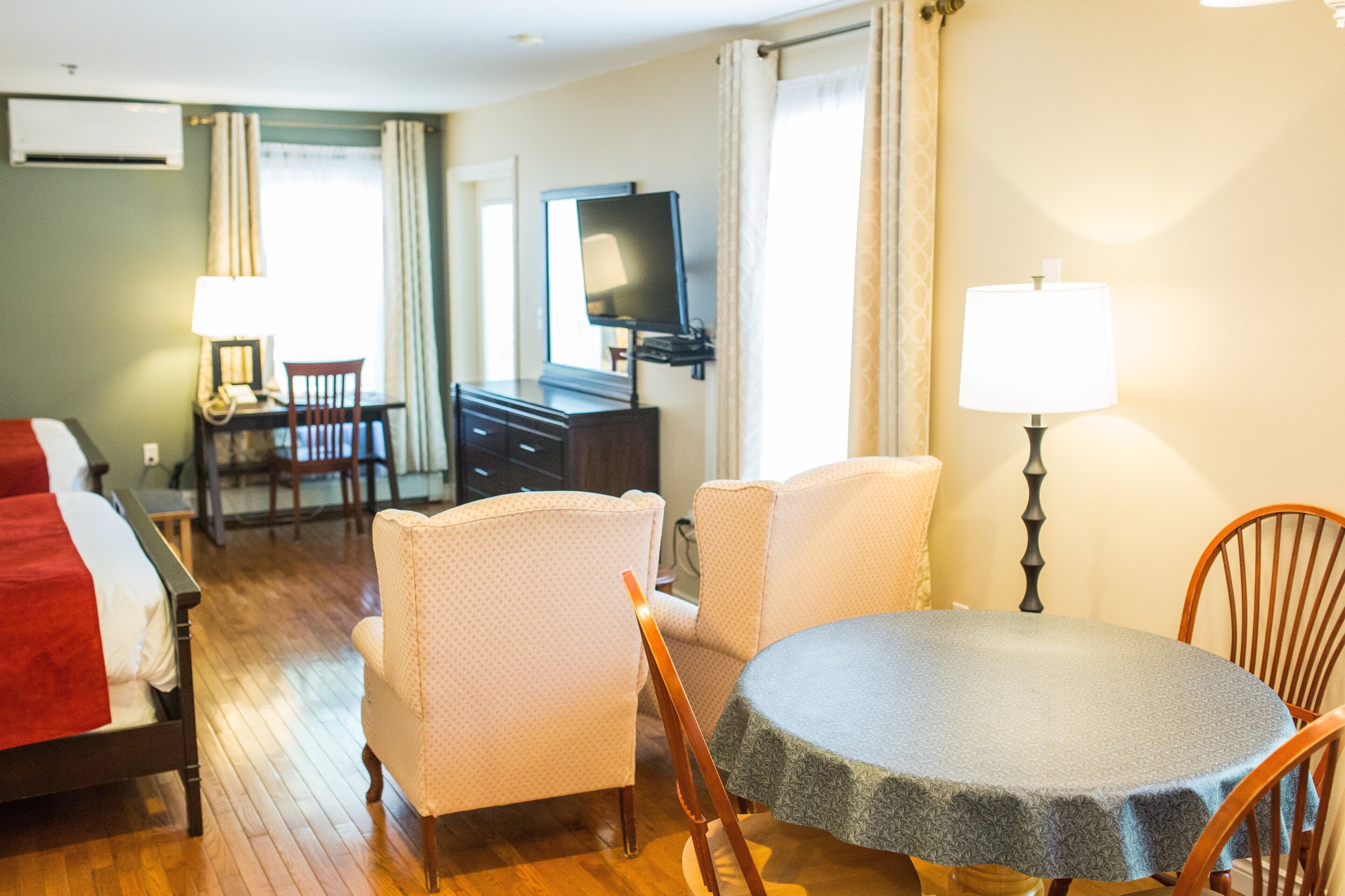 A hotel room with a bed, a desk, a chair, a table, a TV, and a lamp.