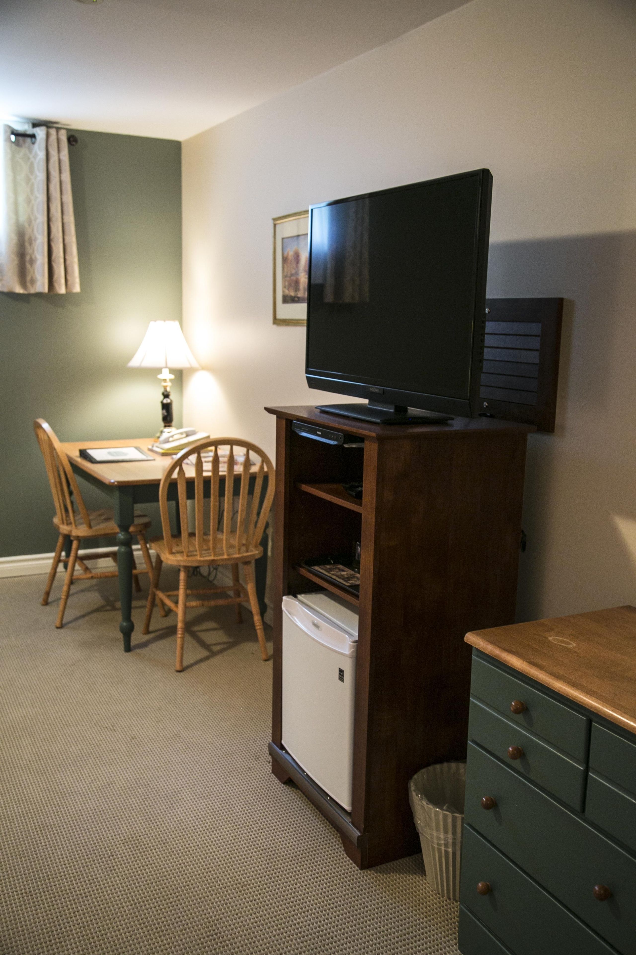 A small room with a television on a wooden stand, a small refrigerator, a wooden chair, a table with a lamp, and a trash can.
