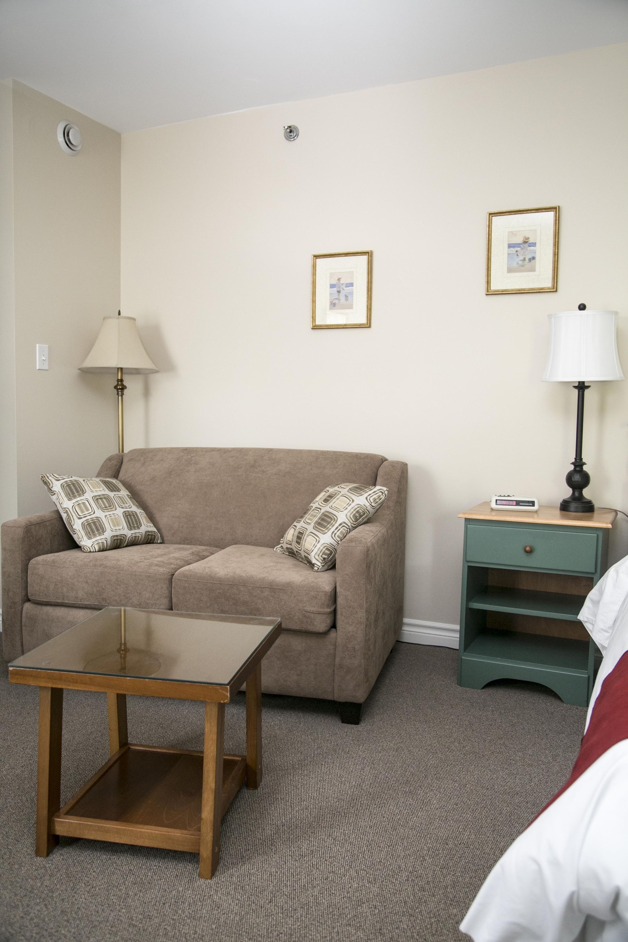 A living room with a beige sofa, two patterned pillows, a glass coffee table, a green nightstand with a lamp, and framed pictures on the wall.