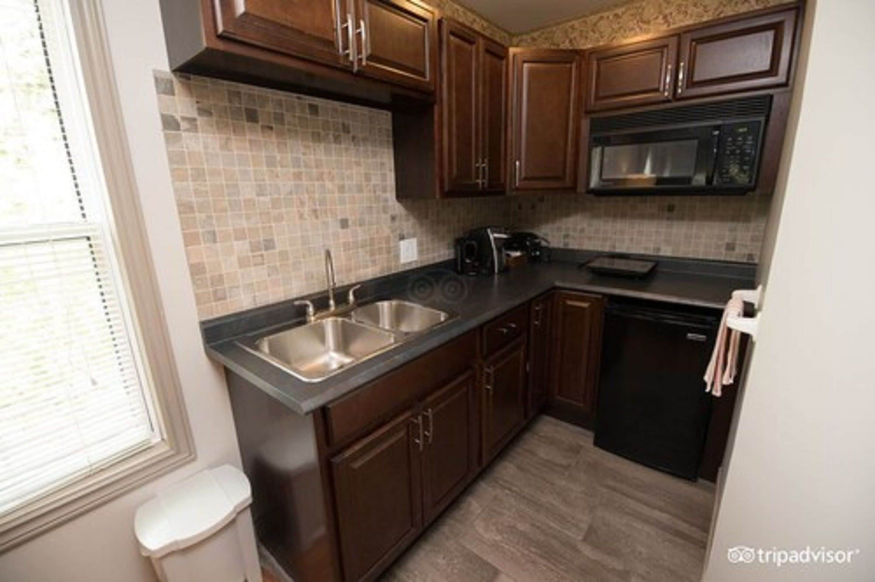 A kitchen with dark wooden cabinets, a tiled backsplash, stainless steel sinks, a microwave, and a dishwasher.