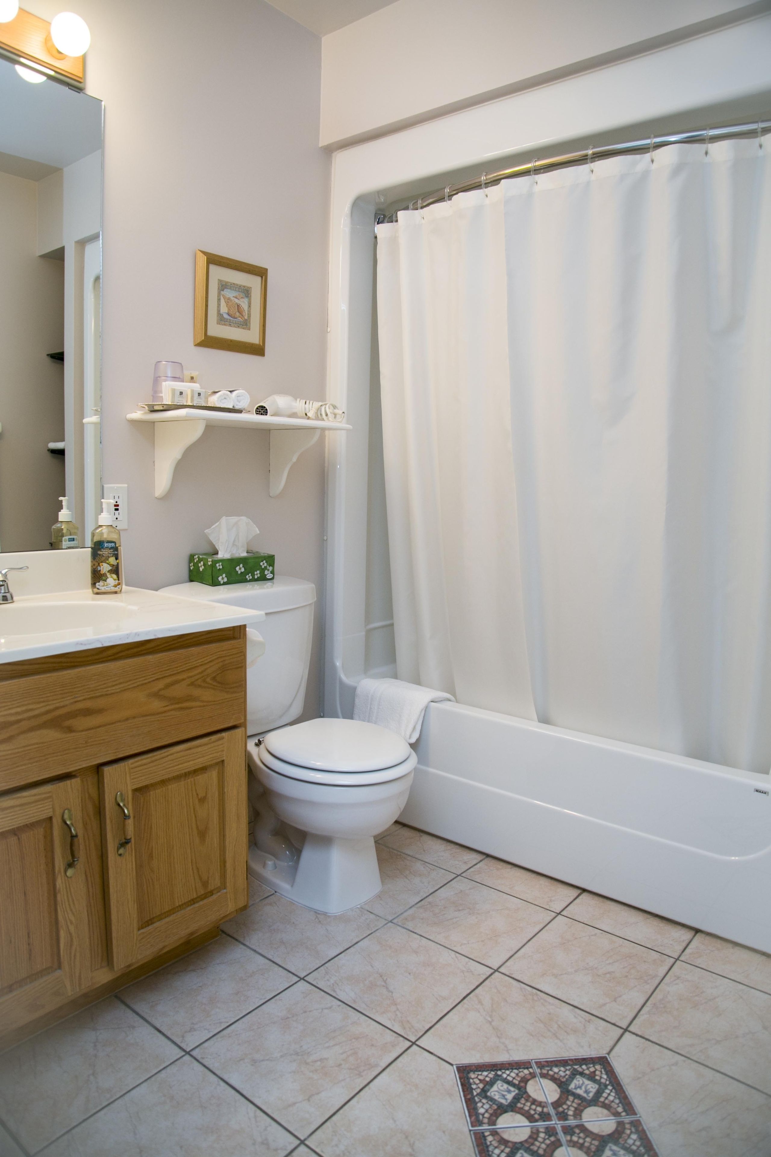 Un baño con inodoro, bañera con cortina de ducha, tocador de madera con espejo y estante con artículos de aseo.