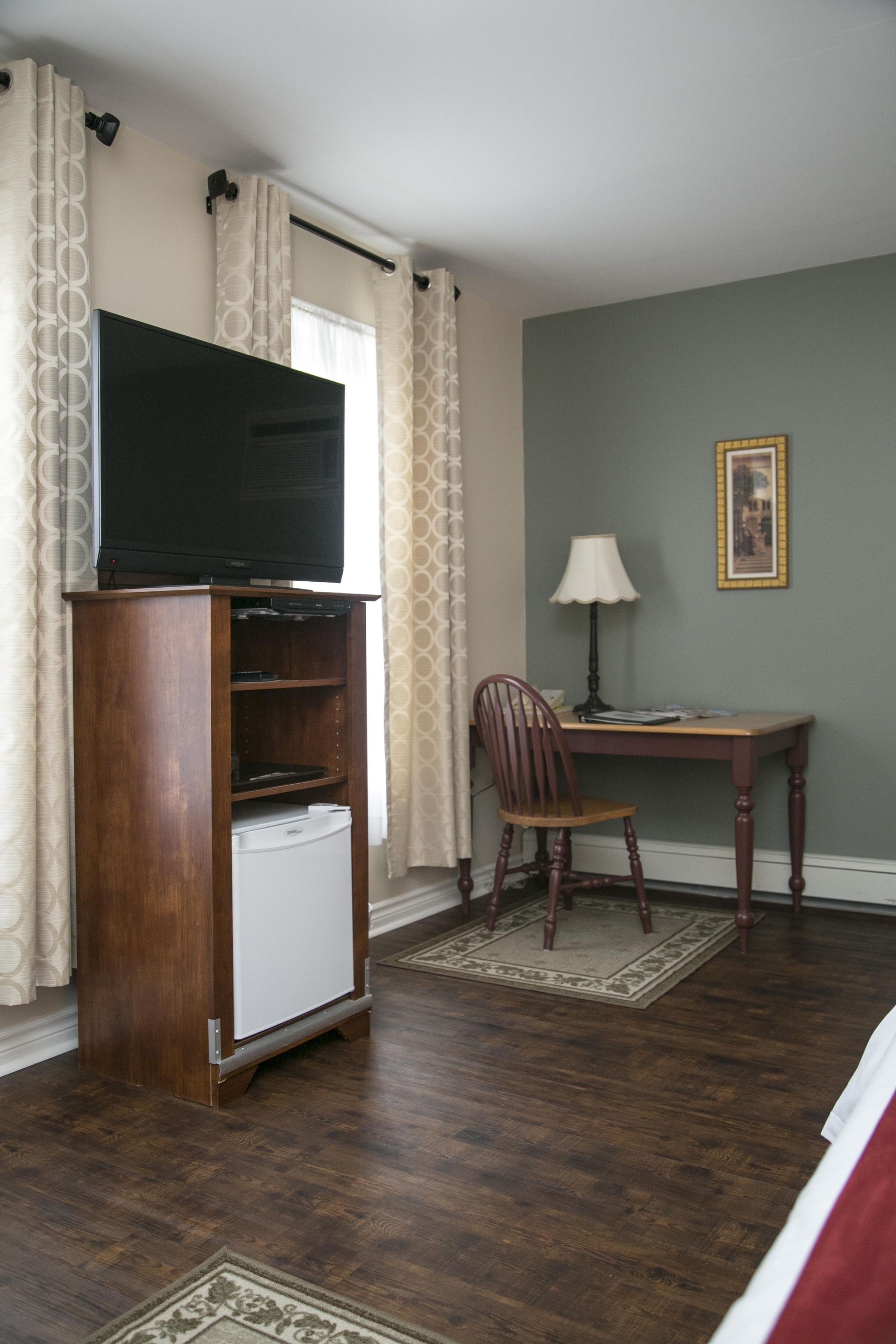 Een kamer met een houten tv-meubel, een bureau met een lamp, een stoel en een lijst met een afbeelding aan de muur.