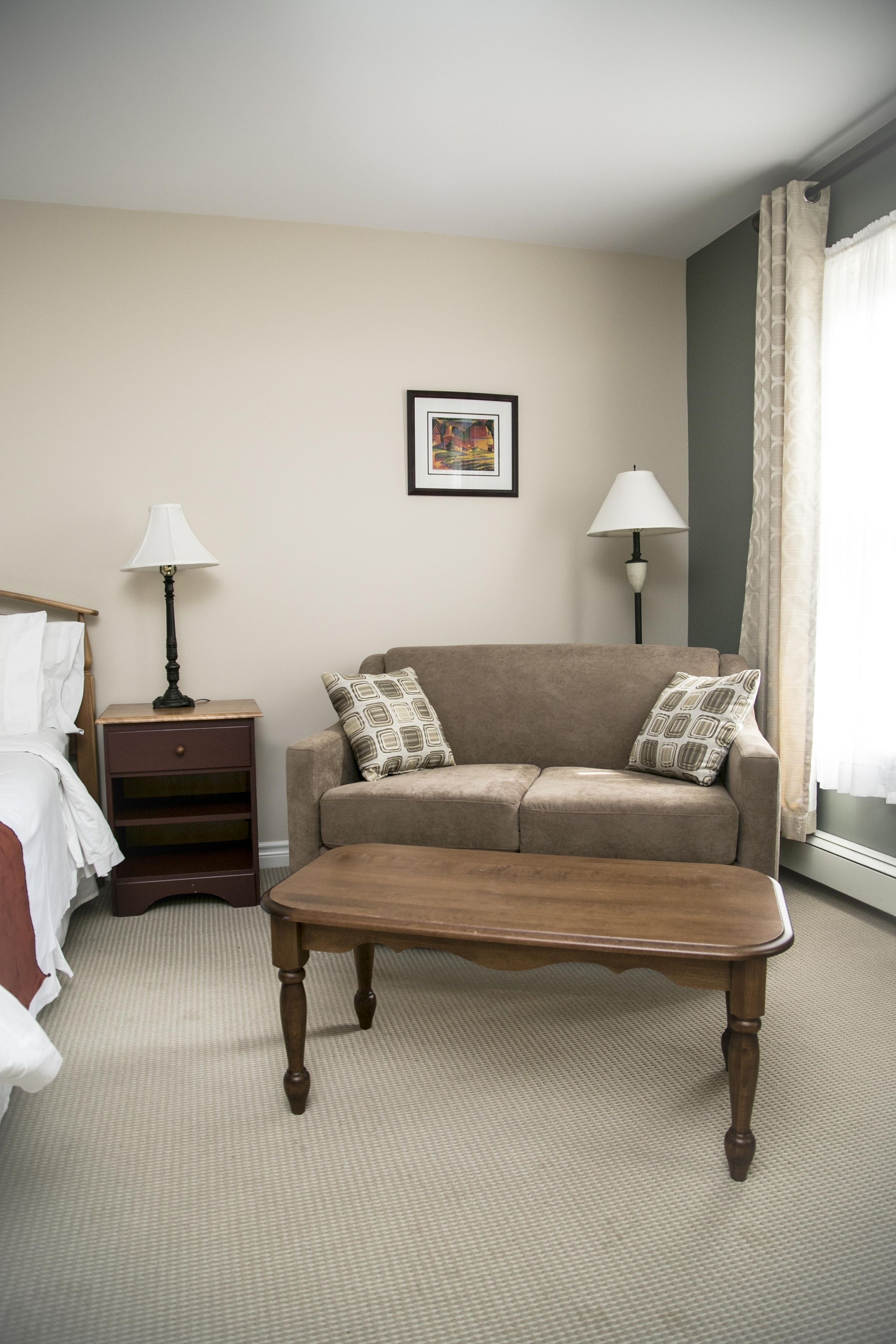 A hotel room with a bed, a wooden nightstand, a beige sofa with patterned cushions, a wooden coffee table, and two wall-mounted lamps.